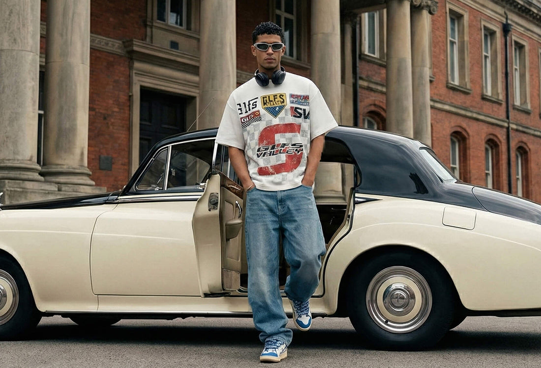A young man wearing sunglasses and headphones leans against the open door of a cream and black vintage luxury car. He is dressed in an oversized white t-shirt with racing-style graphics and baggy blue jeans, standing in front of a grand red brick building with classical stone columns